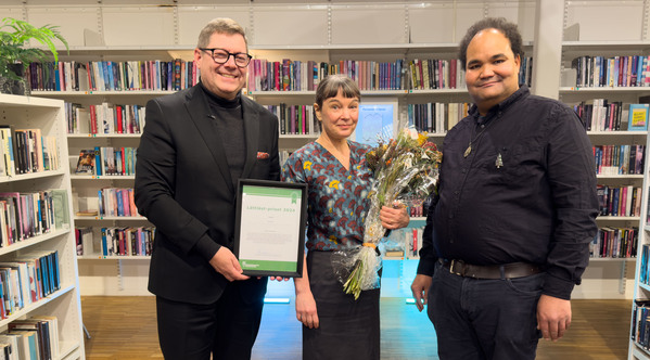 Tre personer står i ett bibliotek, en håller ett diplom och en annan håller en blombukett.