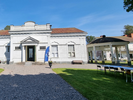 Vit klassisk byggnad med grusgång, grönt gräs och uppställda bord utomhus under en klarblå himmel.