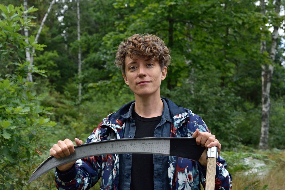 Person står utomhus i skogsmiljö och håller i en lie med båda händerna.