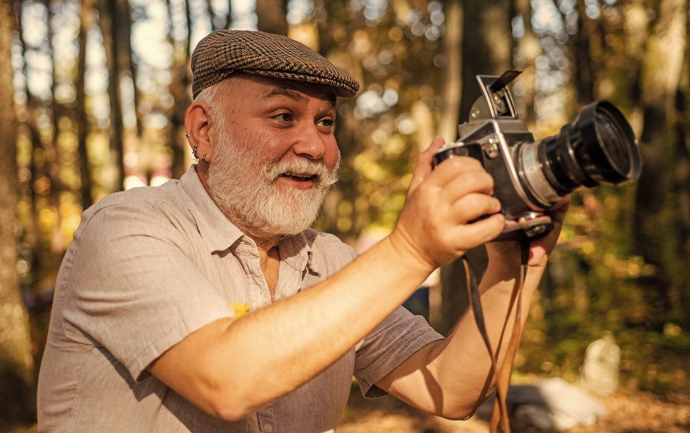 Äldre man med grått skägg och keps fotograferar med en vintagekamera i en solig skog.