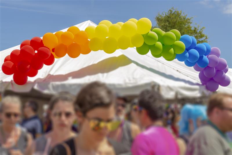 En båge av ballonger i regnbågens färger över ett tält på en utomhusfestival med människor i förgrunden.