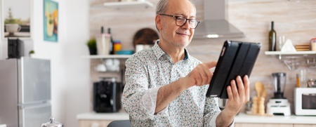 Äldre man sitter vid köksbordet, ler och använder en surfplatta. Frukost med kaffe och frukt på bordet.