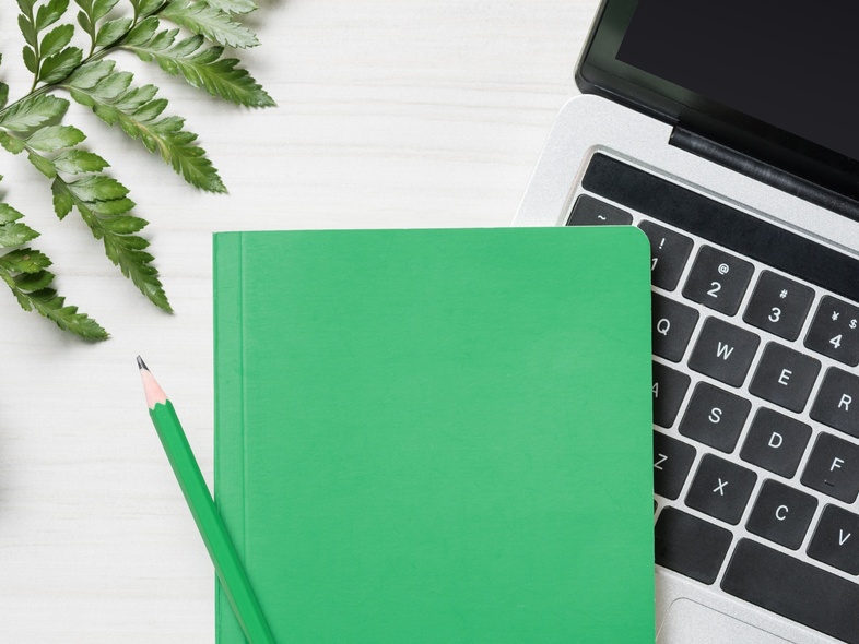 Grön anteckningsbok och penna på en bärbar dator, med växt och ormbunksblad på ett vitt skrivbord.