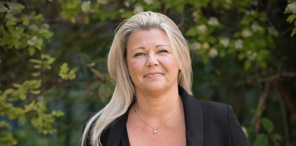 Kvinna med långt blont hår i svart kavaj står utomhus framför gröna buskar.