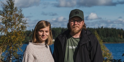 En kvinna och en man står utomhus vid en sjö med skog i bakgrunden under en solig dag.