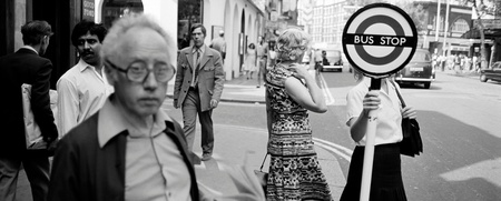 Svartvit gatubild med flera personer vid en busshållplats, en kvinna håller i en busshållplatsskylt.