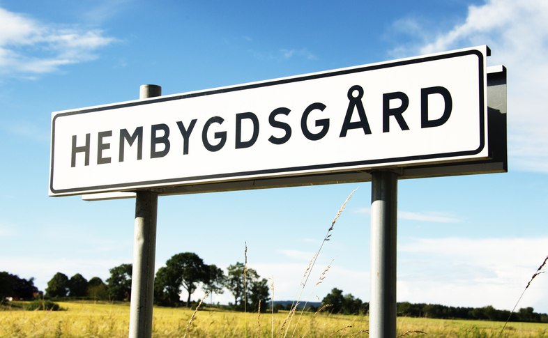 Vägskylt med texten 'Hembygdsgård' mot en lantlig bakgrund med blå himmel och fält.