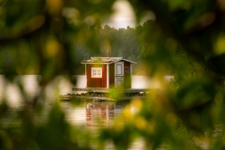 Ett litet rött hus på en brygga vid en sjö, omgiven av suddiga gröna trädgrenar i förgrunden.