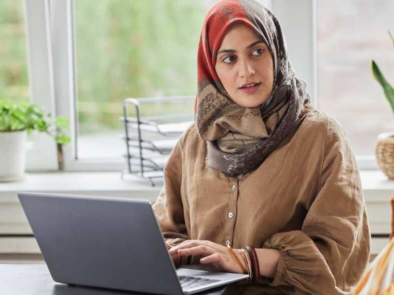 Kvinna framför ett fönster sitter och arbetar vid en laptop.