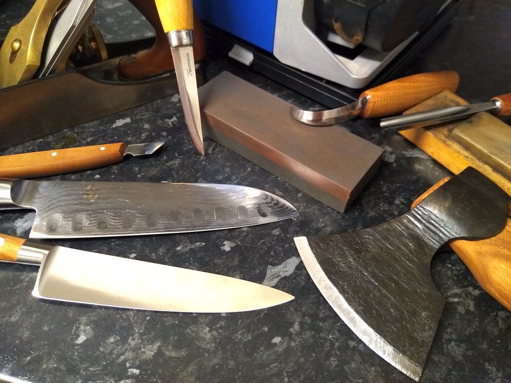 Flera köksknivar, en yxa och ett bryne ligger på en mörk bänkskiva.