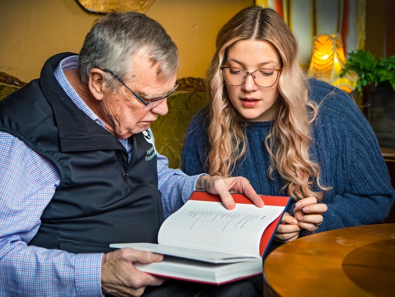 En äldre man och en yngre kvinna sitter tillsammans och läser i en bok i ett vardagsrum.