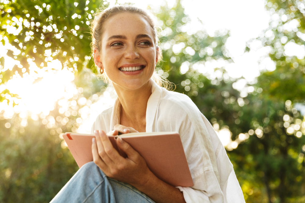 Leende kvinna sitter utomhus med en anteckningsbok i händerna, omgiven av grönska och solljus.