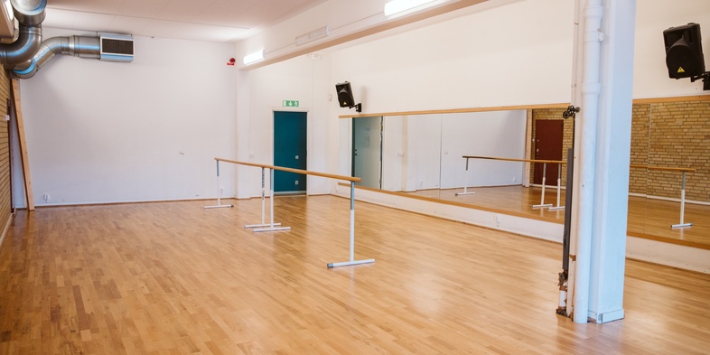 Danssal med trägolv, spegelvägg, balettstång och vita väggar, tom på människor.