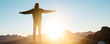 Person står med utsträckta armar på ett berg i soluppgång eller solnedgång.