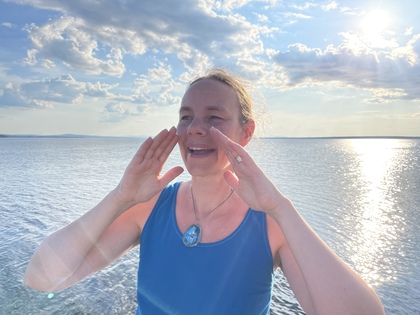 Person i blå linne står vid en sjö med händerna upp, solnedgång och molnig himmel i bakgrunden.