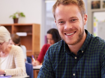 Leende man sitter vid skolbänk i klassrum, skriver i anteckningsbok. Andra vuxna studenter i bakgrunden.