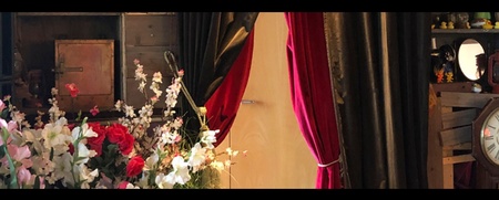 En scen med röda sammetsgardiner, blommor, en klocka och hyllor med föremål i ett varmt ljus.