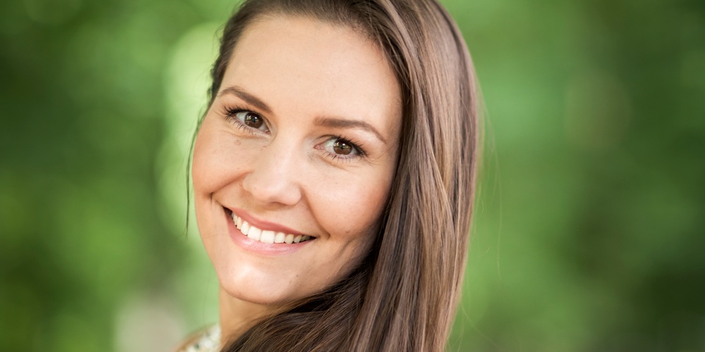 Leende kvinna med långt brunt hår står utomhus mot en suddig grön bakgrund.