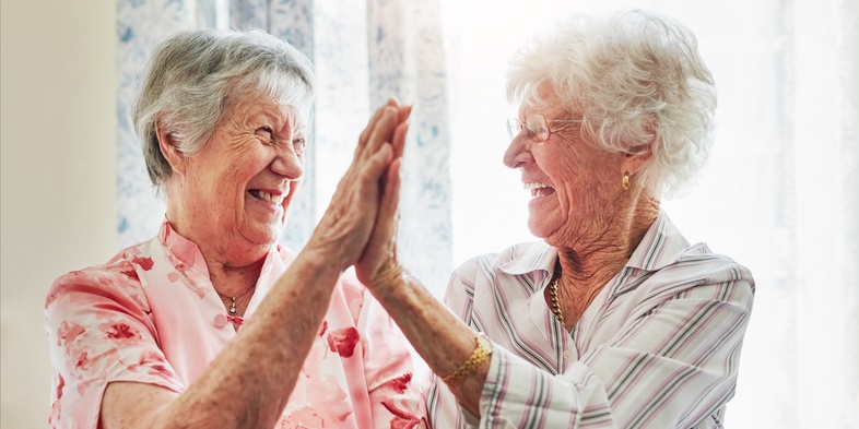Två äldre kvinnor skrattar och ger varandra en high five i ett ljust rum.