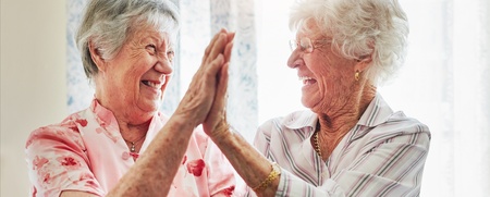 Två äldre kvinnor skrattar och ger varandra en high five i ett ljust rum.