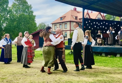 Människor i traditionella folkdräkter dansar utomhus framför en scen med musiker.