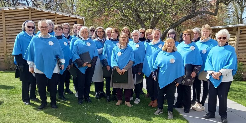 En grupp vuxna står utomhus i blå ponchos, samlade på gräsmatta vid träd och träbyggnad.
