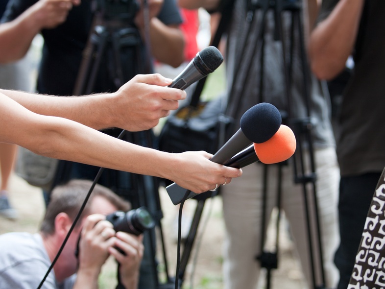 Flera journalister håller fram mikrofoner mot en person under en presskonferens utomhus.