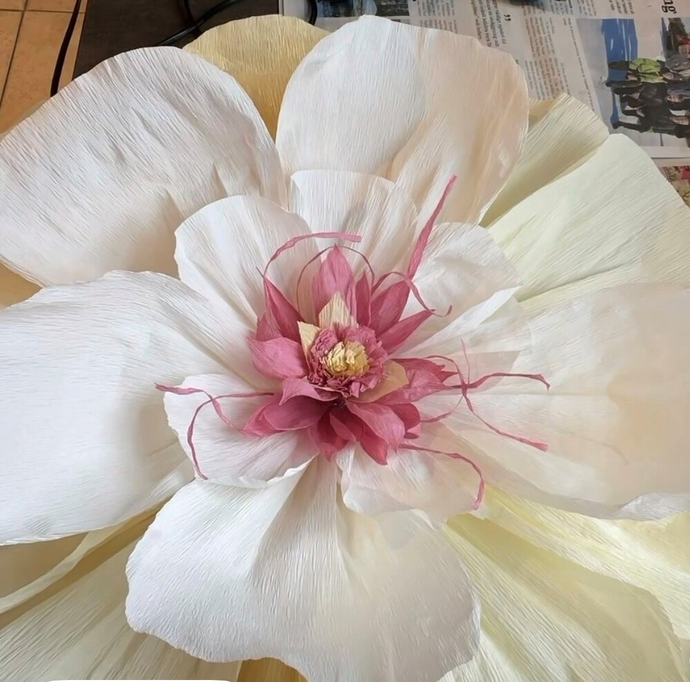 Stor konstgjord blomma av papper med vita och rosa kronblad, liggande på ett bord med tidningar.