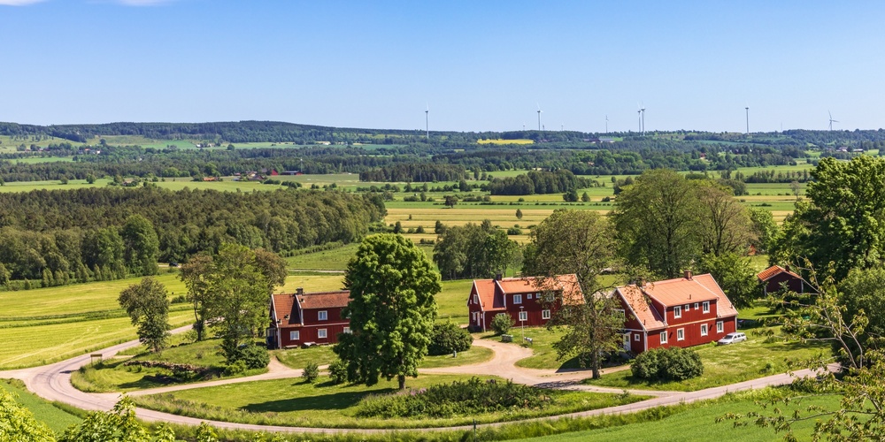 Landskapsvy med röda trähus, gröna fält, träd och vindkraftverk under blå himmel.