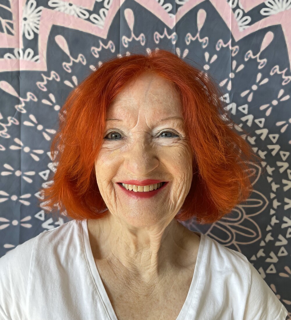 Leende äldre kvinna med rött hår framför ett mönstrat tyg i bakgrunden.