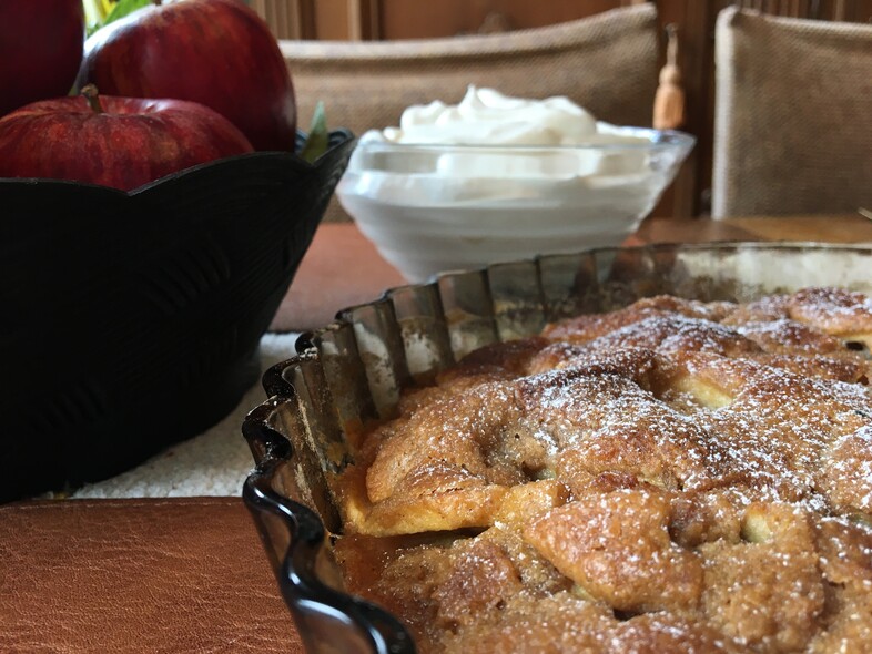 En äppelpaj i en form, en skål med vispgrädde och röda äpplen i en skål på ett bord.