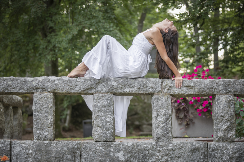 Kvinna i vit klänning poserar liggande på ett stenräcke i en grönskande park.