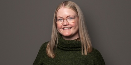 Person med långt blont hår, grön stickad tröja och svarta byxor står mot en grå bakgrund.