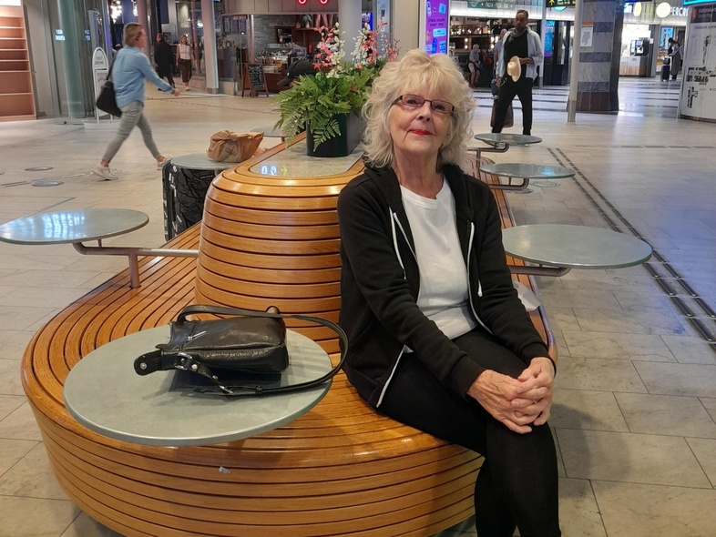 Kvinna sitter på en rund träbänk med små bord i en modern inomhusgalleria med butiker och skyltar.
