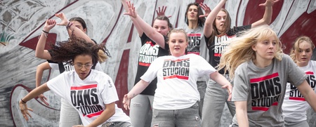 En grupp unga kvinnor dansar energiskt utomhus framför en graffitivägg, alla bär t-shirts med texten 'DANS STUDIO'.