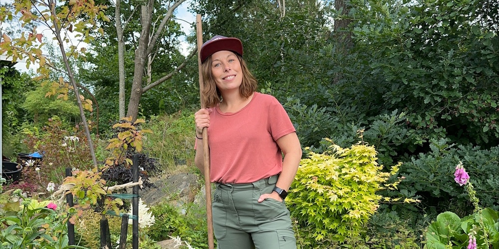 Person står i en trädgård med en röd kratta, omgiven av grönska och blommor.