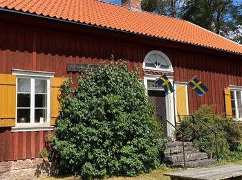 Röd trävilla med gula fönsterluckor, svenska flaggor och picknickbord i trädgård, solig dag.