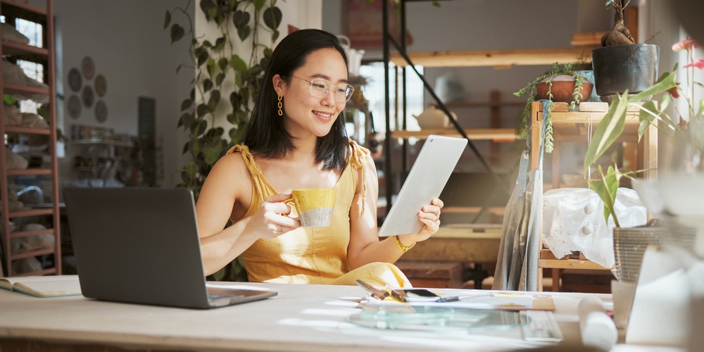 Kvinna i gul klänning sitter vid skrivbord, håller surfplatta och kopp, arbetar i ljust hemmakontor.