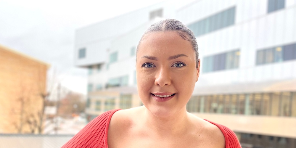 Kvinna i röd tröja står på en balkong med moderna byggnader i bakgrunden.