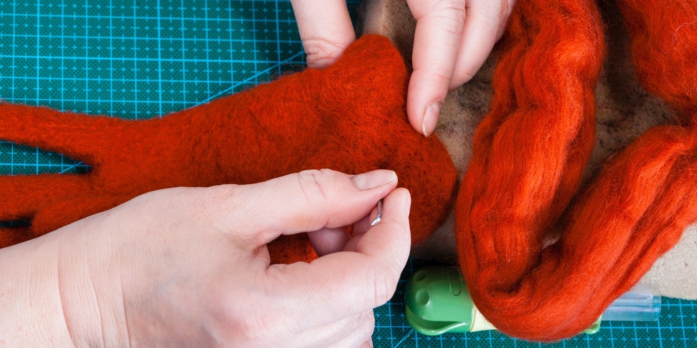 Två händer arbetar med röd ull på en grön skärmatta, troligen för tovning eller hantverk.