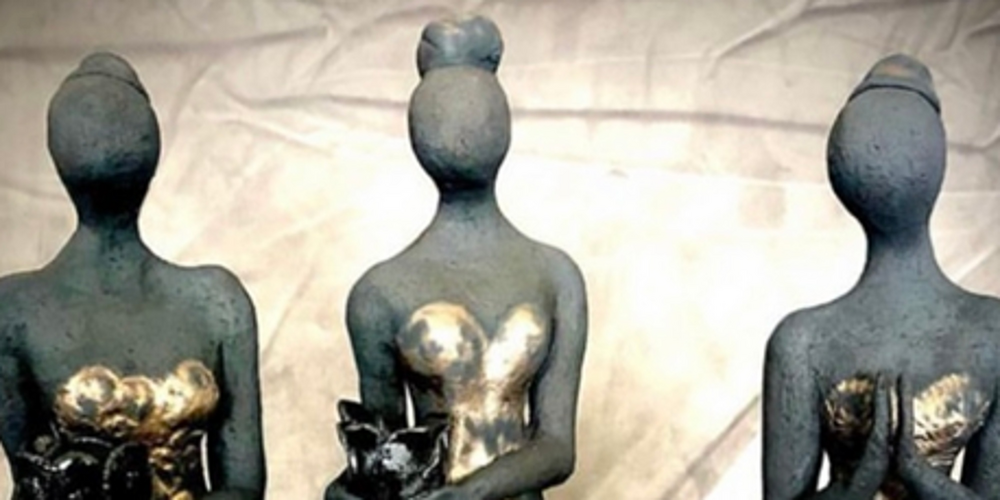 Tre stiliserade kvinnliga statyetter i långa klänningar, utan ansikten, står på rad mot en neutral bakgrund.