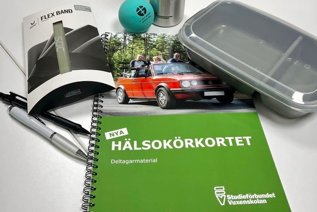 Ett grönt häfte med texten 'Hälsokörkortet', pennor, matlåda, träningsband och broschyr på ett bord.