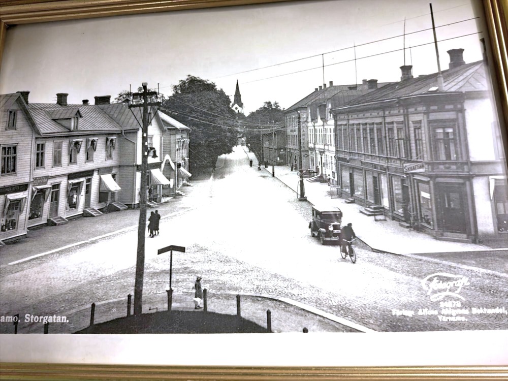Svartvit bild av en stadsgata med gamla trähus, bil, cyklist och några gående personer.