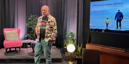 Man står på en scen med svart bakgrund, bredvid en TV-skärm med text och en bild på två personer vid havet.