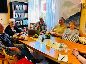 En grupp äldre personer sitter runt ett bord i ett ljust rum, en person står och pratar.