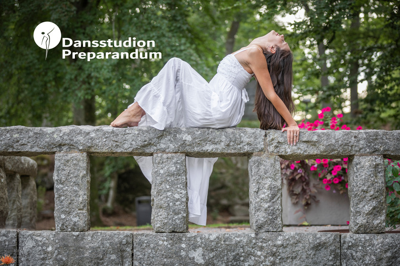 Kvinna i vit klänning poserar dansande på en stenmur utomhus med grönska och blommor i bakgrunden.