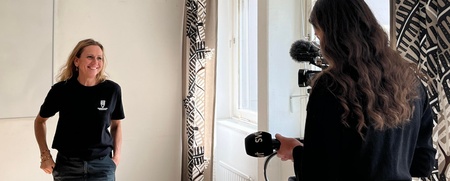 En kvinna står och ler mot en annan kvinna som filmar henne med en kamera i ett ljust rum med mönstrade gardiner.