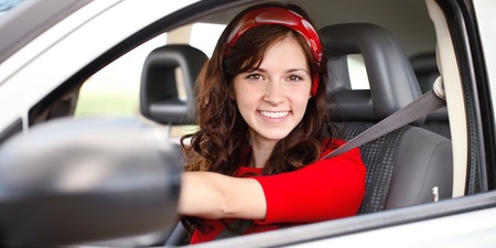 Ung kvinna med långt brunt hår och röd tröja sitter leende bakom ratten i en bil.