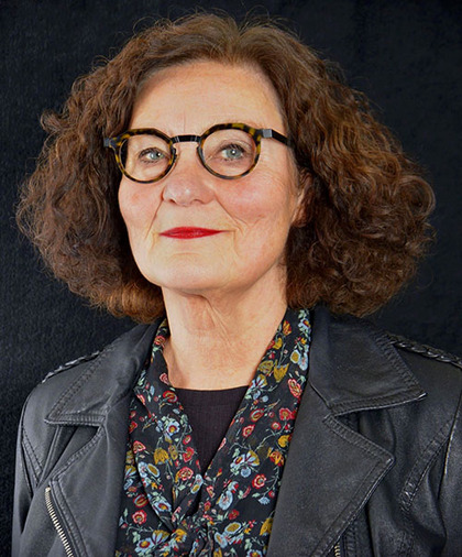 Person med lockigt brunt hår, svart skinnjacka och blommig skjorta mot svart bakgrund.