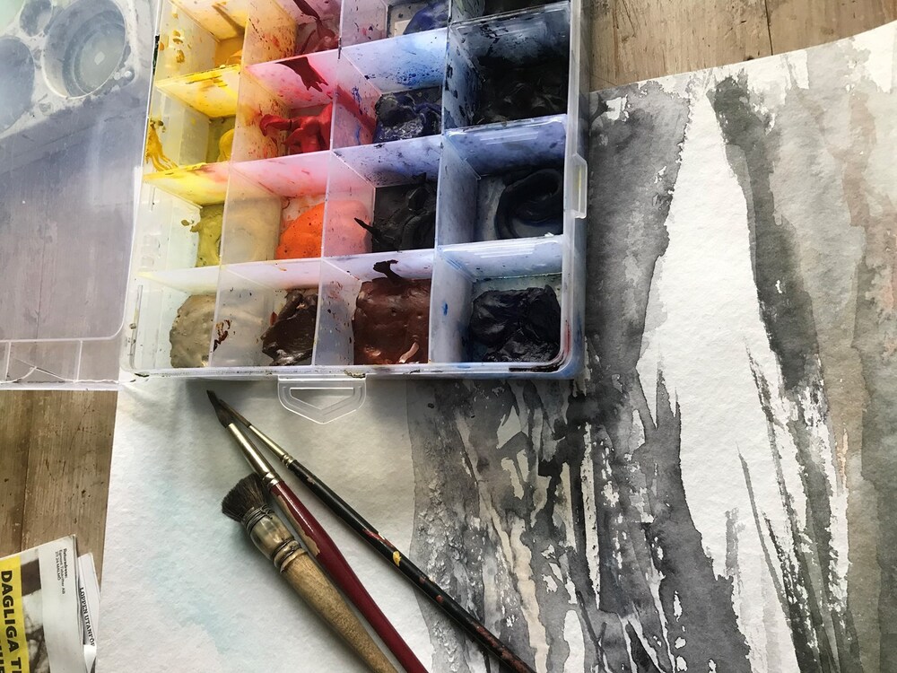 Akvarellfärger, penslar och ett påbörjat konstverk på ett bord.